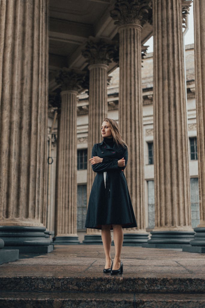 Portrait of a woman in a black coat standing amid classical columns.