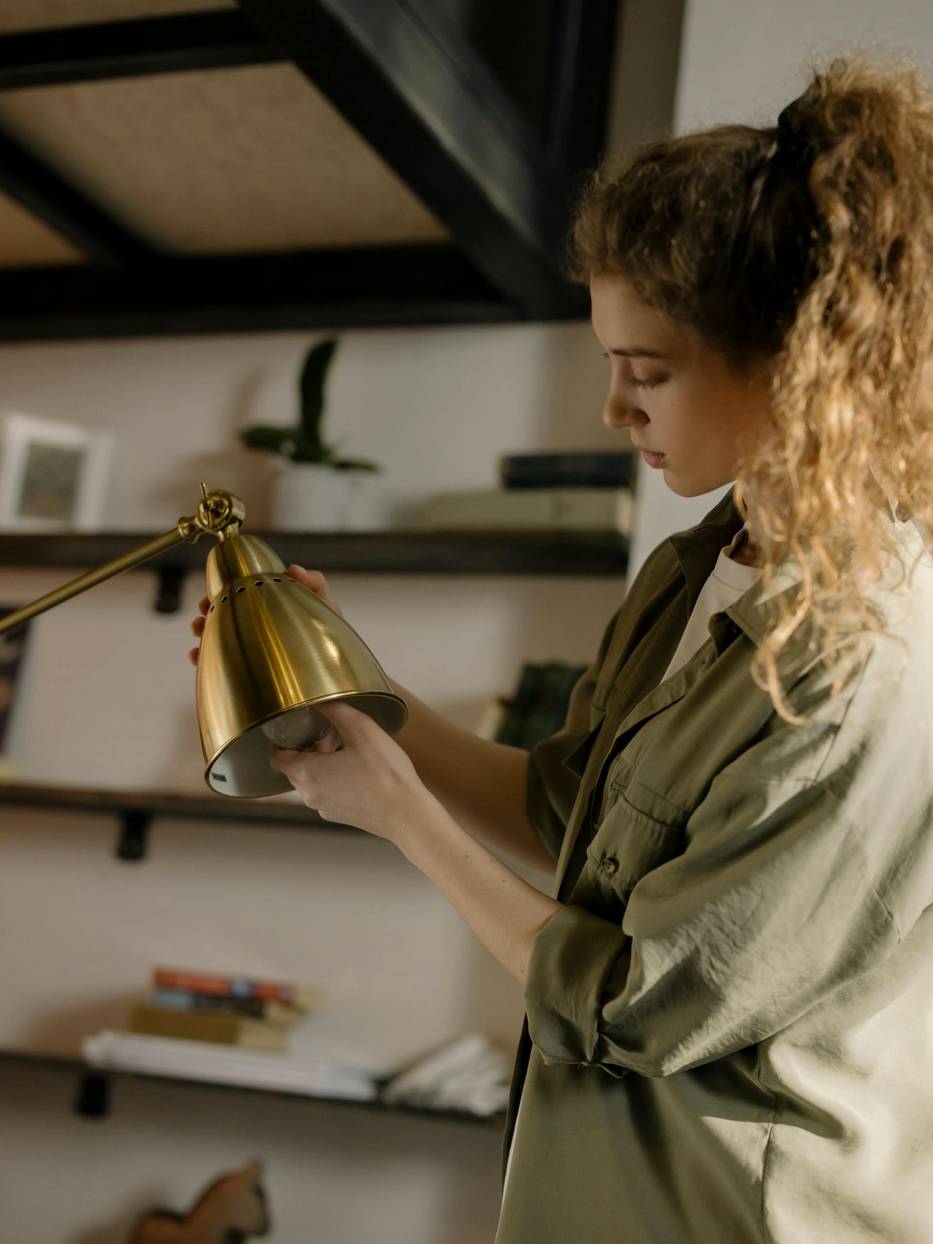 Woman adjusting a golden desk lamp in a cozy home setting with shelves.